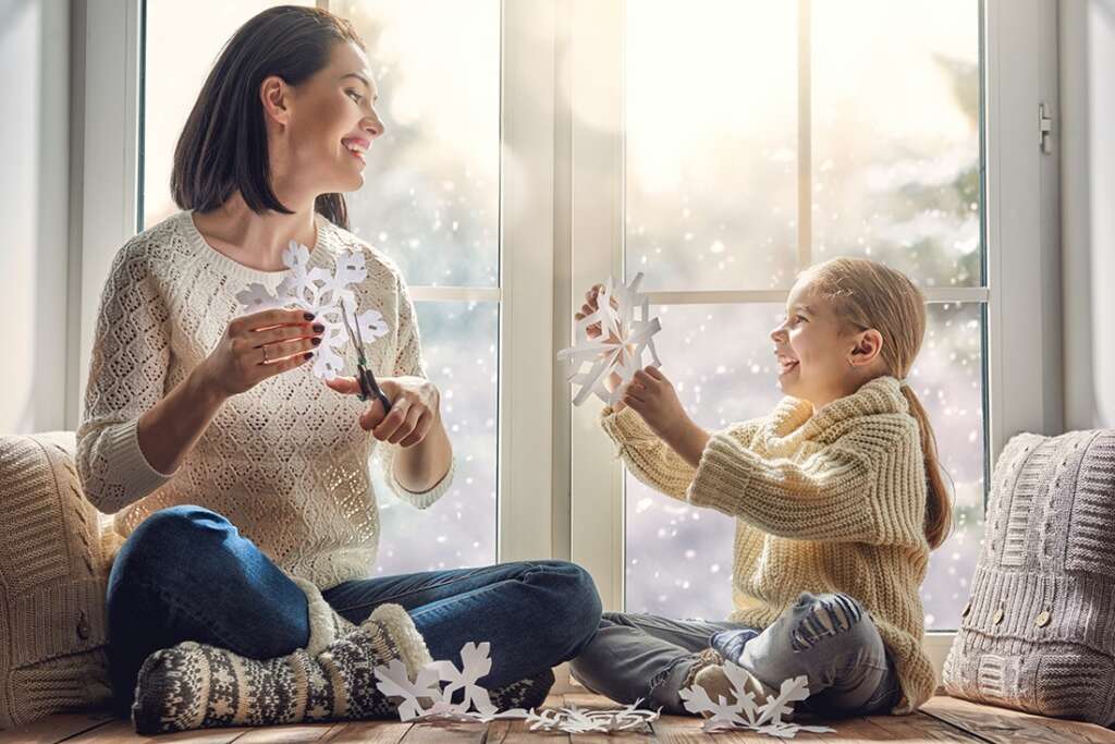 Woman and little girl make paper snowflakes in front of a snowy window